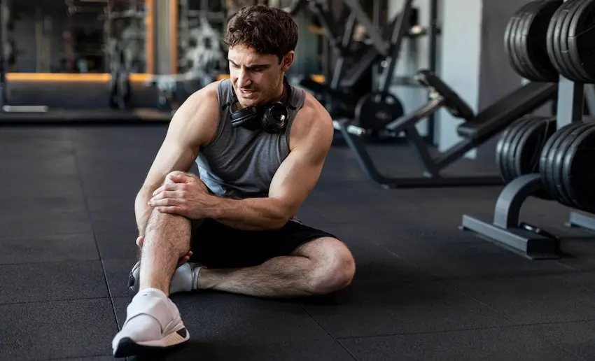 A muscular man in a tank top and headphones sits on a gym floor grimacing while clutching his knee beside a rack of dumbbells representing the type of negligence-related gym injury that may qualify as a claim under personal injury law in Georgia.