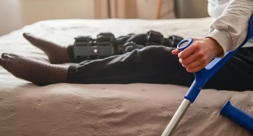 A person wearing a knee brace lies on a bed beside a blue crutch resting against the mattress representing the prolonged physical recovery and lost mobility that personal injury law is designed to help injured victims seek compensation for in Georgia.