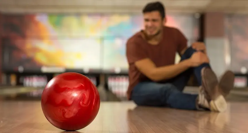 A man in a burgundy shirt sits on a bowling alley lane grimacing in pain while clutching his knee with a red bowling ball resting in the foreground depicting the type of slip and fall injury on a business premises that can lead to a personal injury lawsuit in Tennessee.