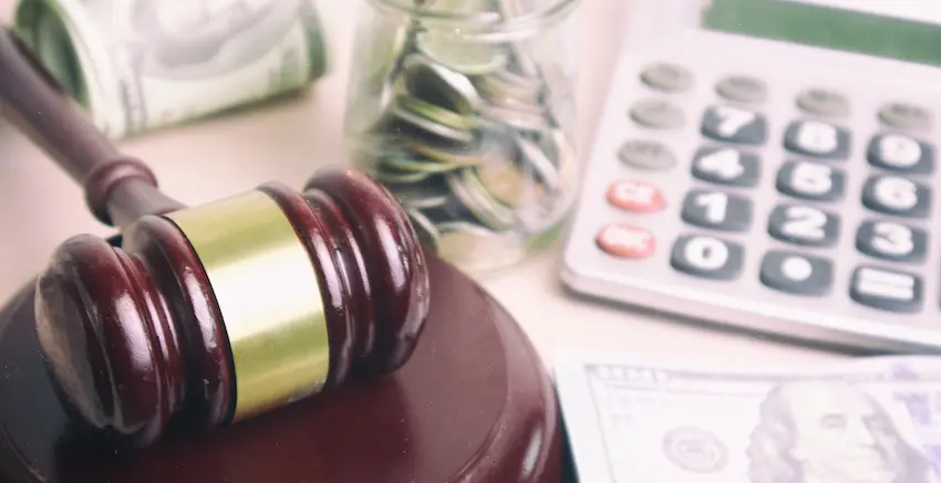 A wooden judge's gavel resting on its sound block beside a glass jar filled with coins- rolled dollar bills- loose hundred-dollar bills- and a calculator on a desk- representing the financial compensation and legal costs involved in pursuing a personal injury lawsuit with Ross Moore Law.