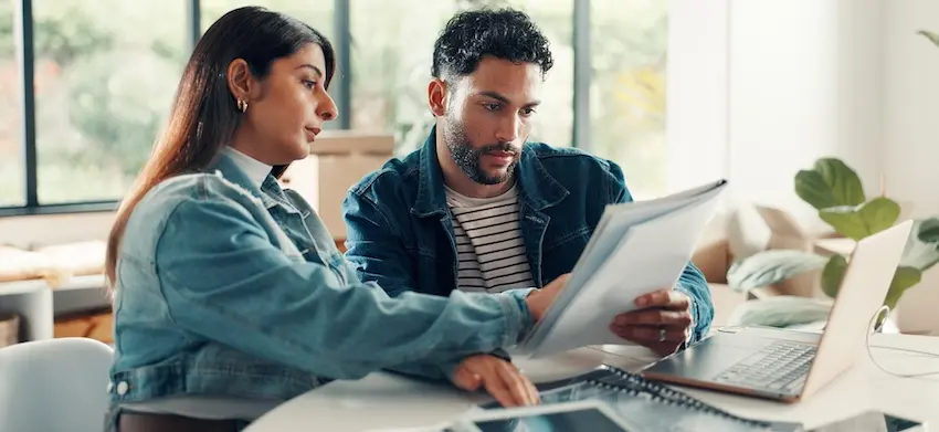 A young woman with long dark hair wearing a light denim jacket leans in closely beside a young man with curly dark hair and a short beard dressed in a teal denim jacket over a striped shirt- both seated at a white table and looking intently at a document he is holding up in both hands with an open laptop on the table in front of them- a large window with soft natural light behind them- and a blurred living room with green plants and cushioned furniture in the background — depicting a couple carefully reviewing the details of their uninsured motorist coverage policy together at home with assistance from Ross Moore Law.
