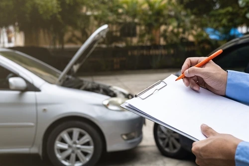 An insurance agent completes a car accident claim form while examining a damaged vehicle.