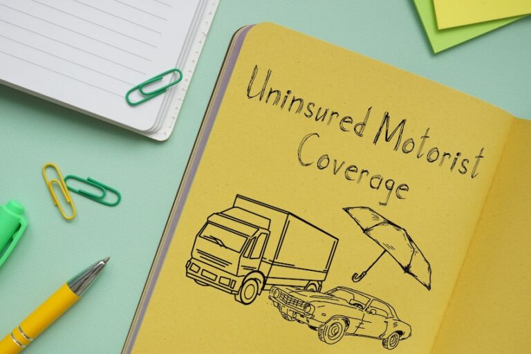 An open yellow-paged notebook lying on a teal surface displays the handwritten phrase Uninsured Motorist Coverage across the top in casual script lettering with a hand-drawn illustration below showing a large commercial box truck looming over a smaller vintage-style sedan alongside an open umbrella symbolizing financial protection while a yellow ballpoint pen- two green paper clips- a yellow paper clip- a lined white notepad- and a small yellow sticky note are scattered around the notebook on the teal surface — a creative visual explanation of what uninsured motorist coverage protects drivers against in Georgia.