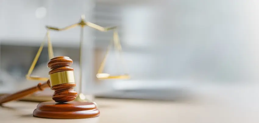 A polished warm-toned wooden judge's gavel with a wide gold band sits upright on its matching circular sound block on a light beige surface in the left foreground while a large ornate brass balance scale with two evenly suspended pans stands slightly blurred in the center background against a softly lit neutral gray office environment — representing the legal process of pursuing justice and fair compensation through an uninsured motorist coverage claim with help from Ross Moore Law in Georgia.