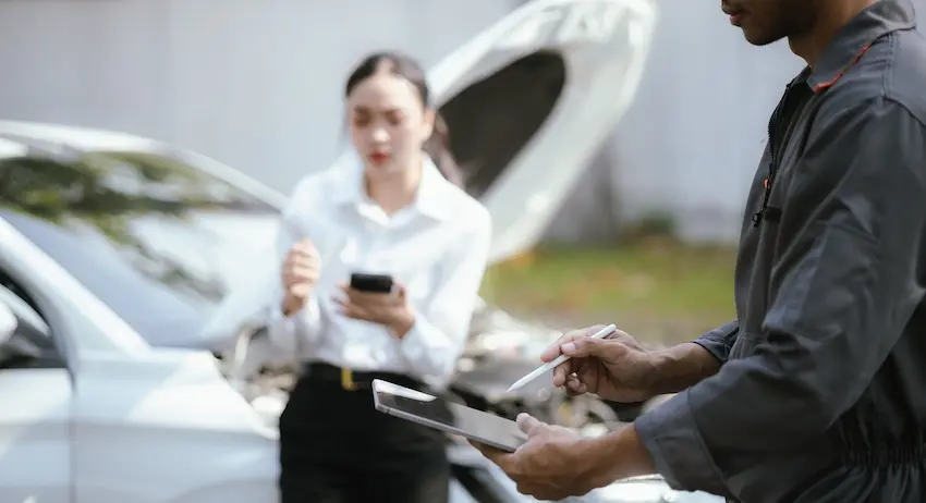 A man in a dark gray mechanic's uniform stands in the right foreground holding a white tablet and a stylus while documenting vehicle damage while a young woman in a white dress shirt and black pants stands in the blurred background leaning against a silver car with its hood propped fully open- looking down at her smartphone with visible front-end damage to the vehicle suggesting a recent collision — illustrating the post-accident documentation and reporting process that typically initiates an uninsured motorist coverage claim in Georgia.