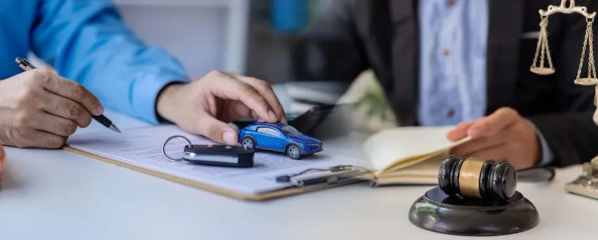 A client and attorney from Ross Moore Law review paperwork on a desk beside a miniature blue car- car keys- a gavel- and a pair of scales of justice- representing the legal consultation process involved in pursuing a car accident lawsuit.