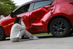 Johns Creek car accident victim sitting beside damaged red vehicle with crushed door after collision.